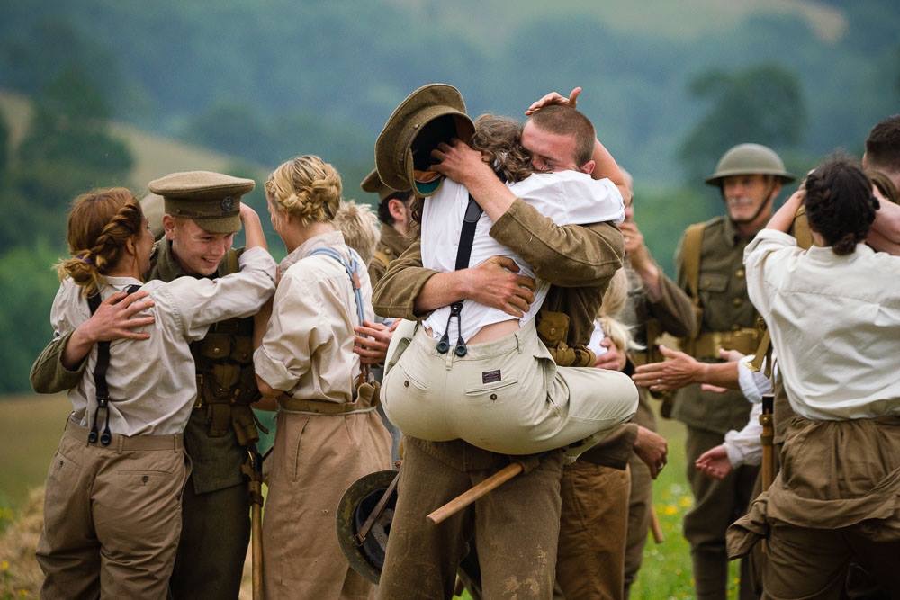 Two WWI-costumed performers embrace in a field as others around them reunite, hills in the background