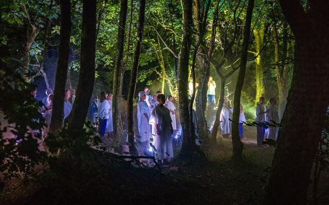 A procession of performers in white moves through a dark woodland lit by scattered lanterns