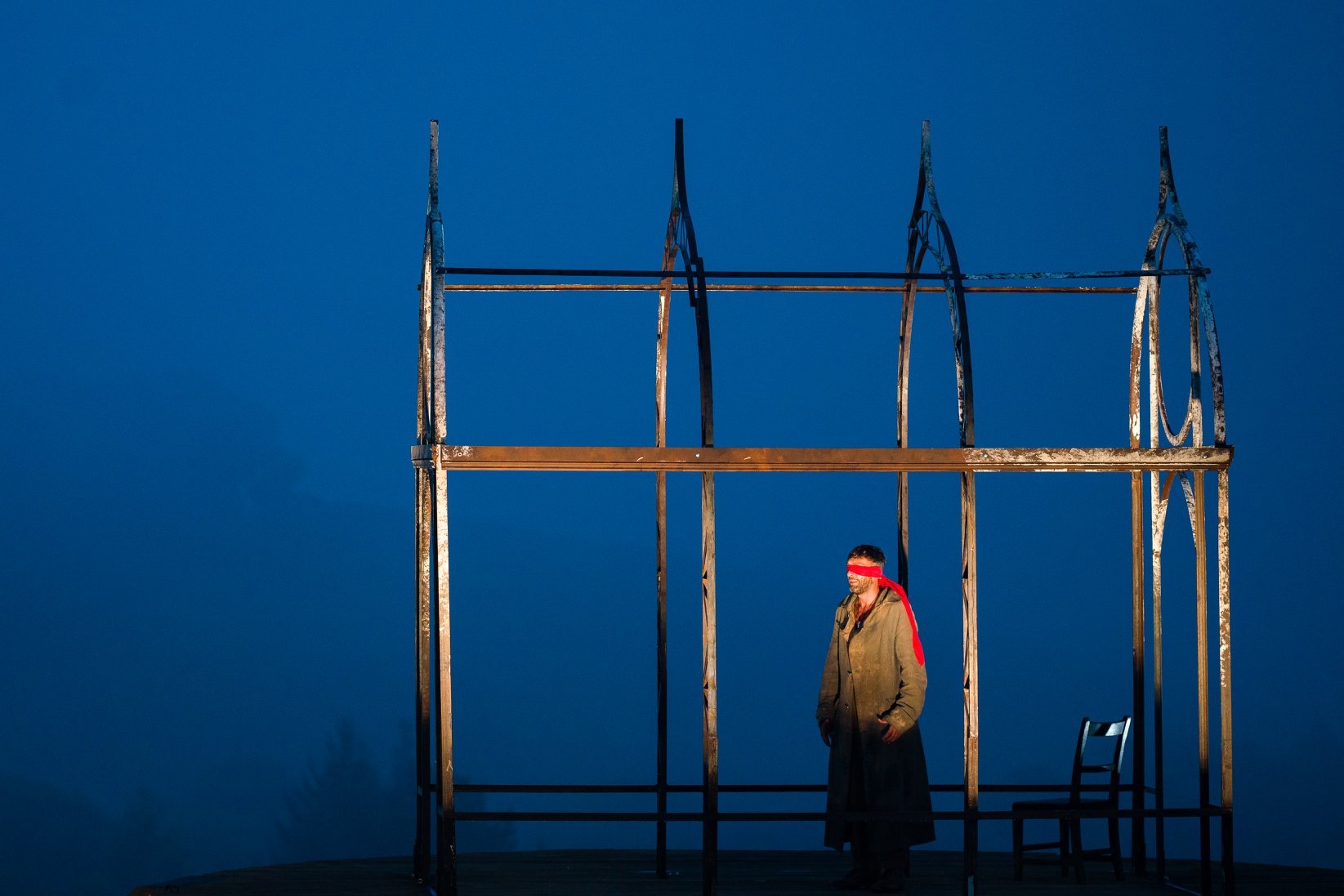 A blindfolded performer in a red blindfold stands beneath a tall rusted metal frame against a deep blue evening sky