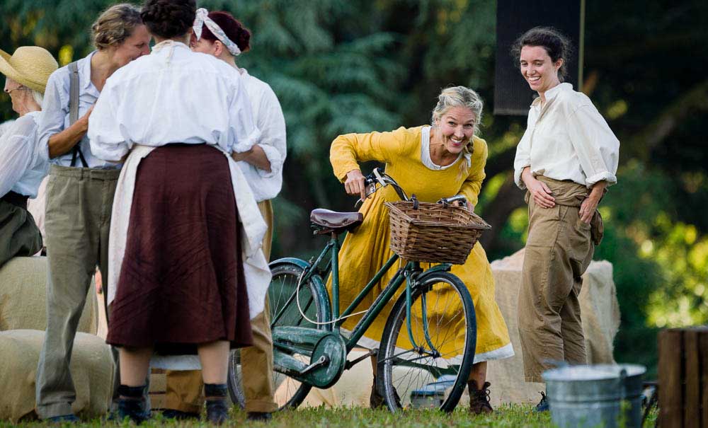 A performer in a yellow dress laughs while wheeling a bicycle with a wicker basket, surrounded by other costumed performers