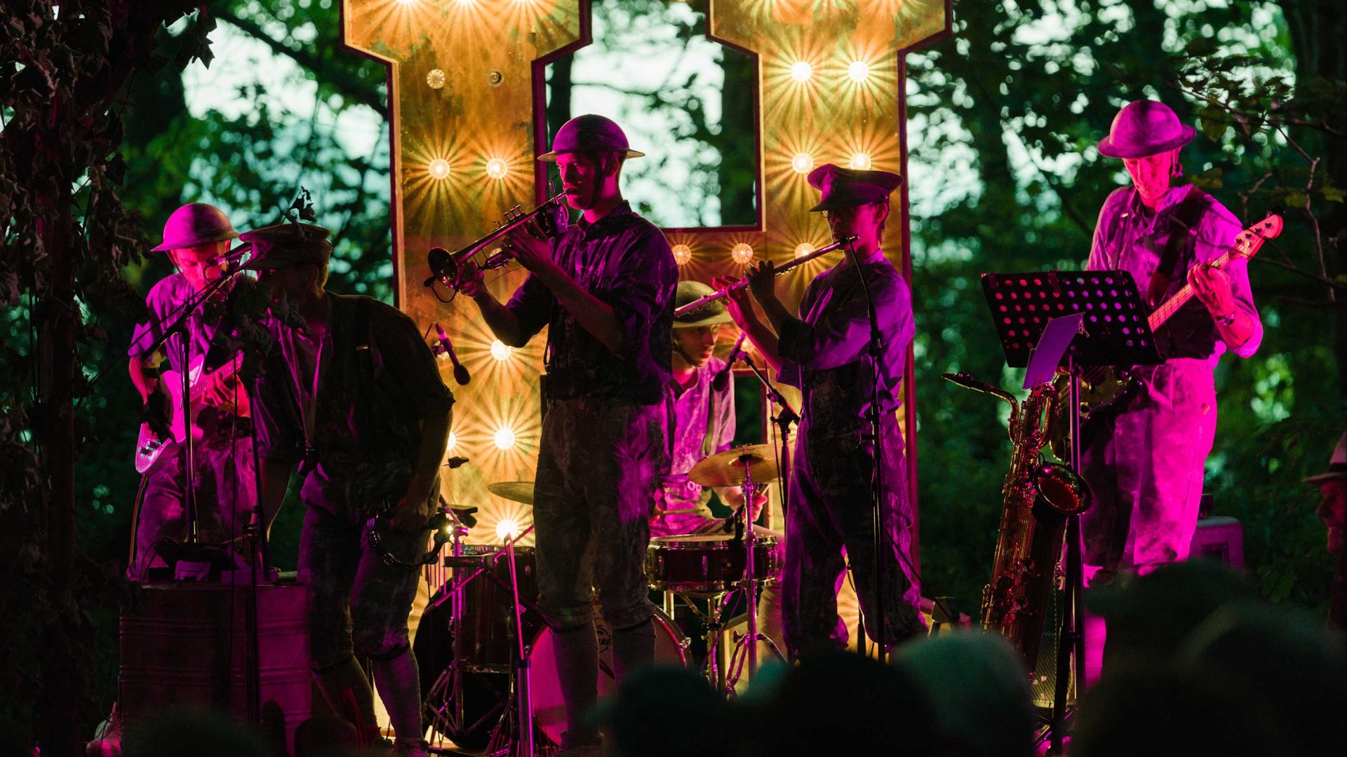 A band in military-style costume plays trumpet, flute, saxophone and guitar on an outdoor stage lit in pink and gold