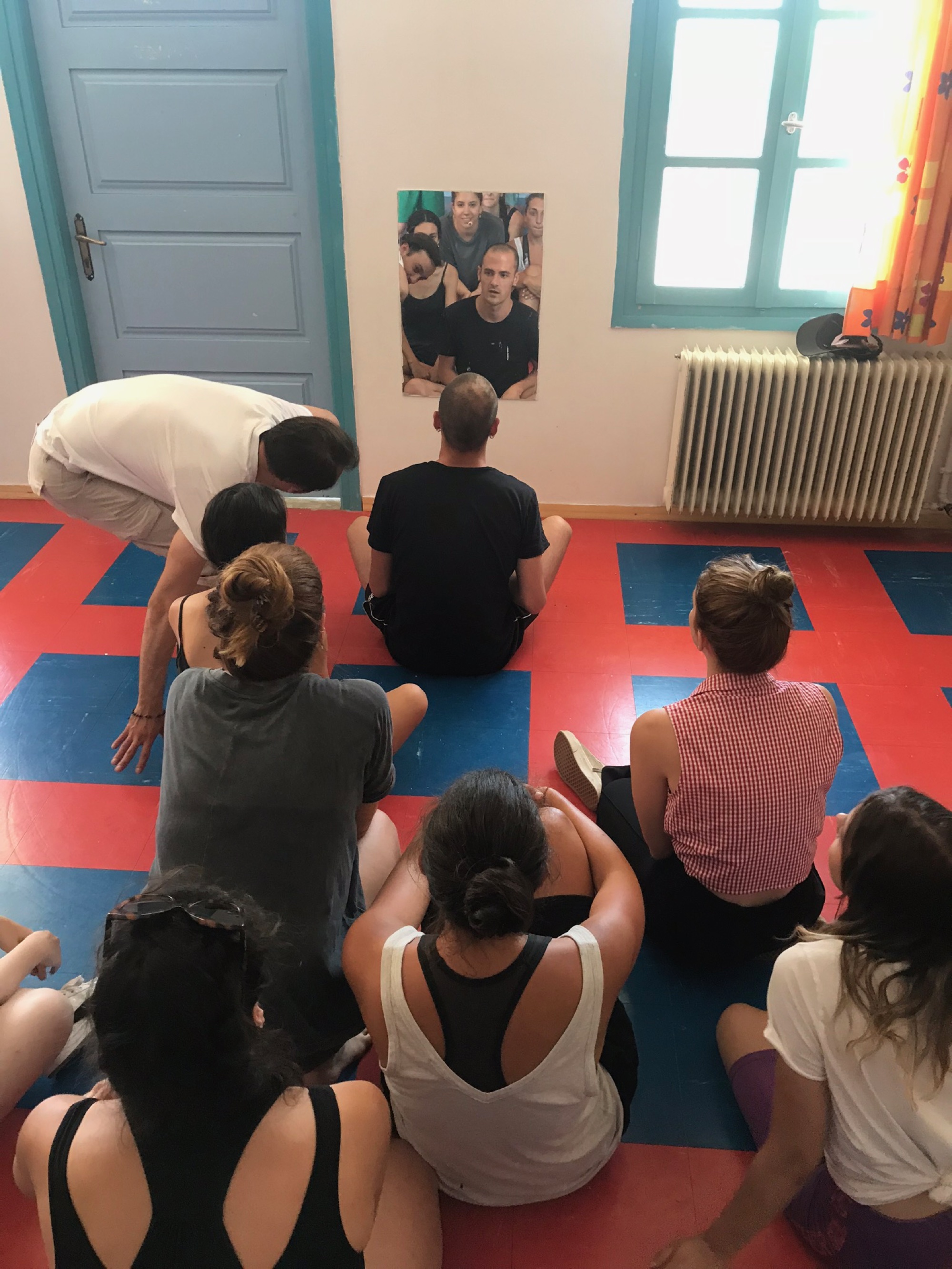A performer sits cross-legged before a photograph of a huddled group, surrounded by others seated on a red and blue tiled floor