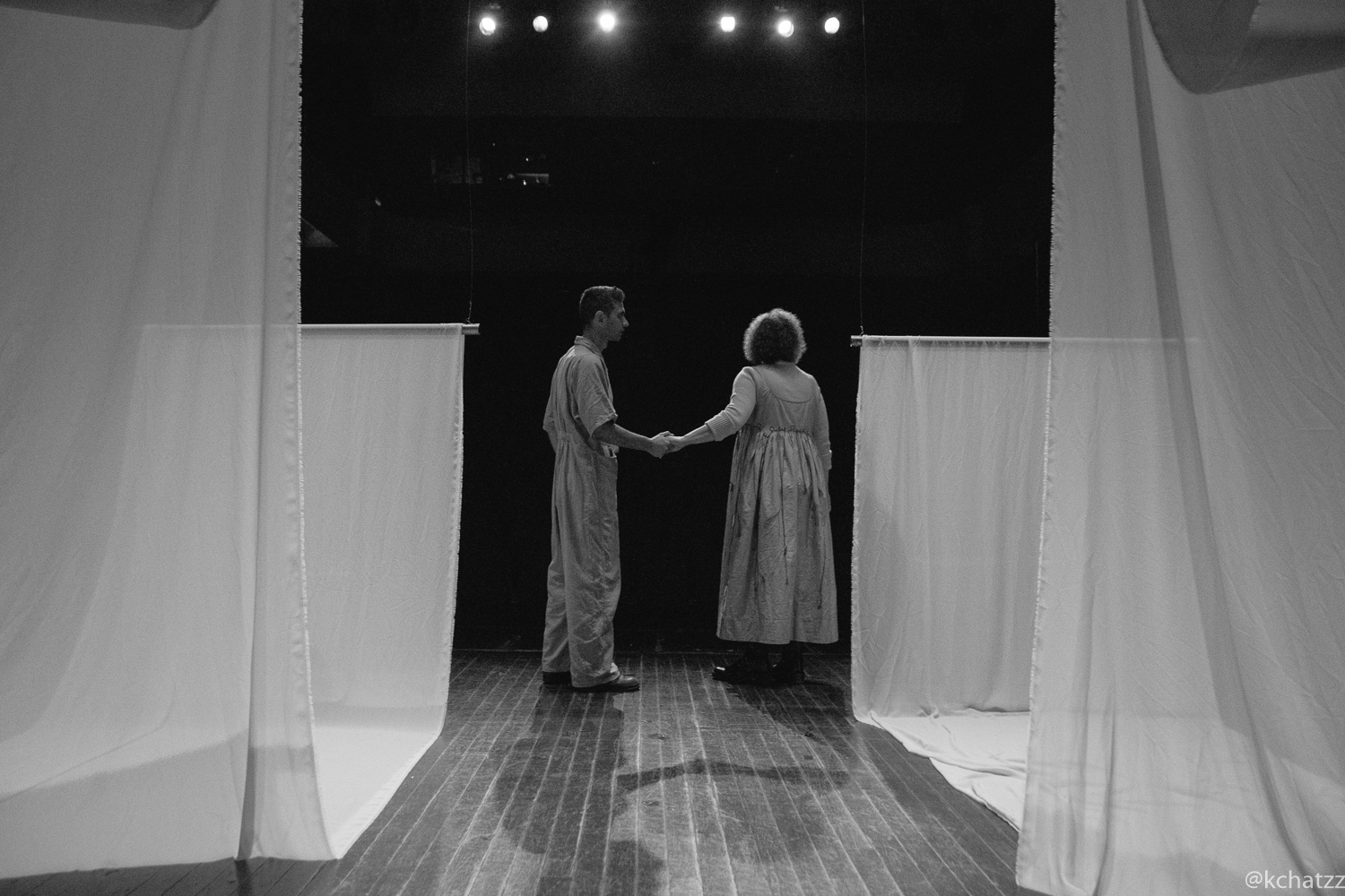 Two performers from Bouboulina: Untold hold hands behind their backs on a bare stage lit by overhead stage lights