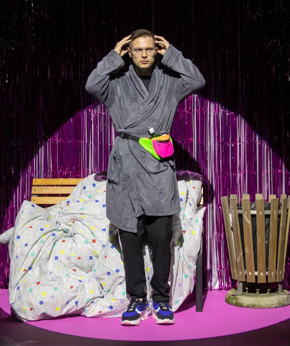A performer in a dressing gown and a neon bum bag stands on stage against a pink tinsel backdrop for First Time