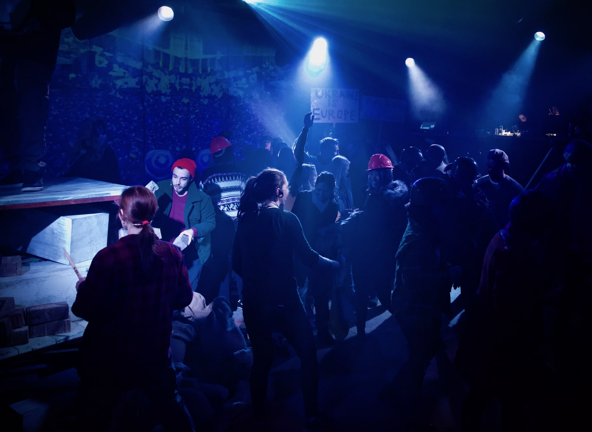 A crowd of performers and audience members in red hard hats under blue stage light, one holding a sign reading Ukraine is Europe
