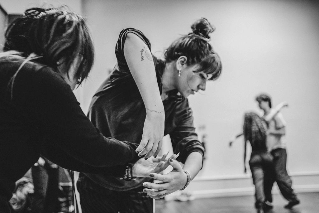 A performer bends forward, hands clasped near her chest as if embracing someone unseen, others close beside her